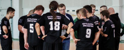 Drużyna w czarnych strojach z numerami 5, 9, 18, 14, 2 i 1 zbiera się na hali sportowej. Kontekst: mecz lub trening.