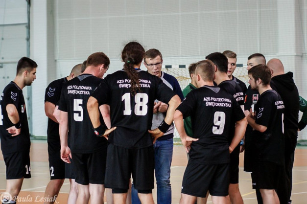 Drużyna w czarnych strojach z numerami 5, 9, 18, 14, 2 i 1 zbiera się na hali sportowej. Kontekst: mecz lub trening.