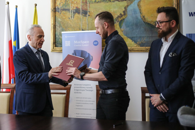 Trzech mężczyzn w garniturach i koszulach. Jeden z nich przekazuje dokument drugiemu. W tle flagi i obraz.