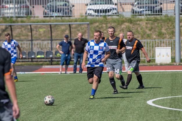 Piłkarze w strojach sportowych biegną po boisku. W tle ławki rezerwowych i samochody.