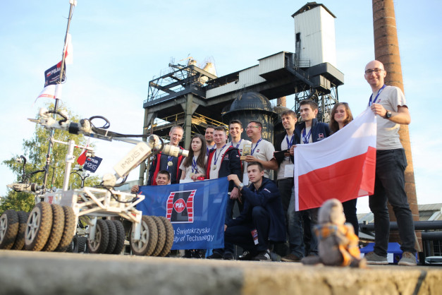 Grupa ludzi z polską flagą i robotem na tle przemysłowej zabudowy. Kluczowe obiekty: robot, ludzie, flaga Polski, budynki.