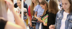 Grupa młodych ludzi ogląda materiały informacyjne na targach edukacyjnych. Kluczowe obiekty: broszury, młodzi ludzie.
