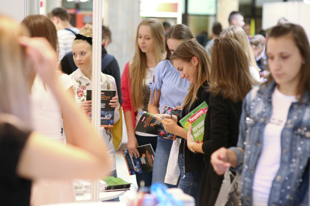 Grupa młodych ludzi ogląda materiały informacyjne na targach edukacyjnych. Kluczowe obiekty: broszury, młodzi ludzie.