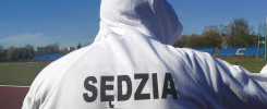 Osoba w białej bluzie z kapturem z napisem "SĘDZIA" stoi na bieżni stadionu. W tle widać trybuny i boisko.