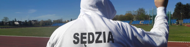 Osoba w białej bluzie z kapturem z napisem "SĘDZIA" stoi na bieżni stadionu. W tle widać trybuny i boisko.