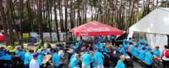 Ludzie w niebieskich koszulkach siedzą i stoją pod czerwonym parasolem Coca-Cola. Kontekst: wydarzenie plenerowe.