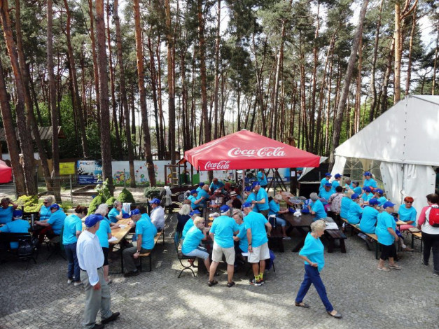 Ludzie w niebieskich koszulkach siedzą i stoją pod czerwonym parasolem Coca-Cola. Kontekst: wydarzenie plenerowe.