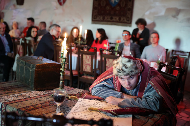 Mężczyzna w stroju historycznym pochyla się nad dokumentem. W tle widać ludzi przy stołach, świece i skrzynię.