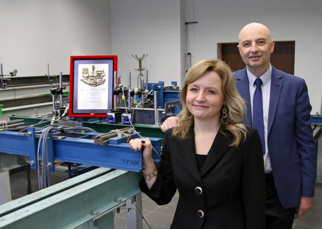 Kobieta i mężczyzna stoją obok siebie w laboratorium. W tle widać ramę z dyplomem i aparaturę badawczą.
