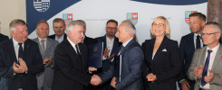 Mężczyźni i kobieta w garniturach stoją przed banerem z herbem.
