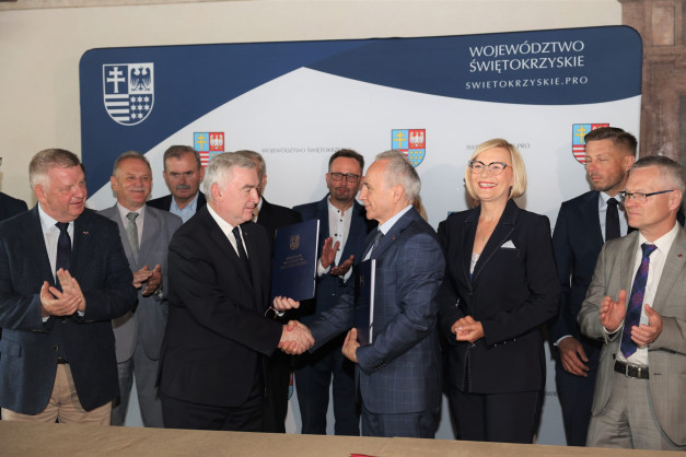 Mężczyźni i kobieta w garniturach stoją przed banerem z herbem.
