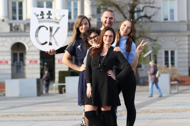 Pięć osób pozuje do zdjęcia z tabliczką "CK" i koroną. W tle budynek i drzewa.
