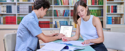 Dwoje studentów uczy się w bibliotece. Kluczowe obiekty: laptop, książki, zeszyty. Kontekst: nauka, biblioteka.