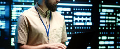 Mężczyzna z brodą pracuje na laptopie w serwerowni. W tle widać rzędy świecących serwerów.