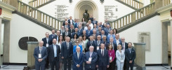 Grupa ludzi pozuje do zdjęcia na schodach. W tle widnieje rzeźba i tablice pamiątkowe.