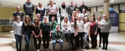 Grupa młodych ludzi pozuje do zdjęcia w holu budynku. W tle widnieje tablica informacyjna.