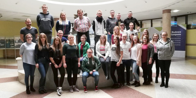 Grupa młodych ludzi pozuje do zdjęcia w holu budynku. W tle widnieje tablica informacyjna.