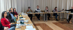 Grupa osób siedzi przy stole, niektórzy noszą okulary VR. Na stole leżą dokumenty i kubki.