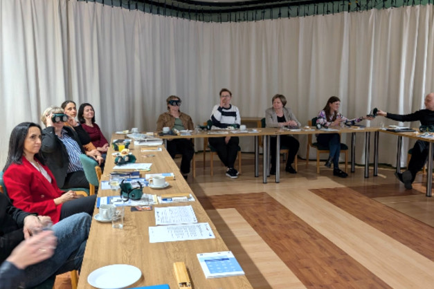 Grupa osób siedzi przy stole, niektórzy noszą okulary VR. Na stole leżą dokumenty i kubki.