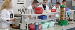 Osoby w fartuchach pracują w laboratorium. Kluczowe obiekty: fartuchy, miski, sita, próbki. Kontekst: laboratorium badawcze.