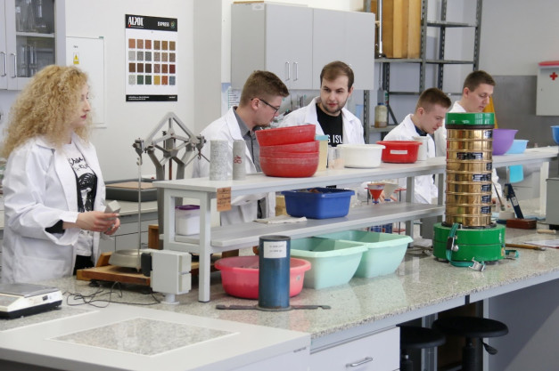 Osoby w fartuchach pracują w laboratorium. Kluczowe obiekty: fartuchy, miski, sita, próbki. Kontekst: laboratorium badawcze.