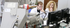 Dwóch naukowców w białych fartuchach pracuje przy skomplikowanym sprzęcie laboratoryjnym i monitorach.