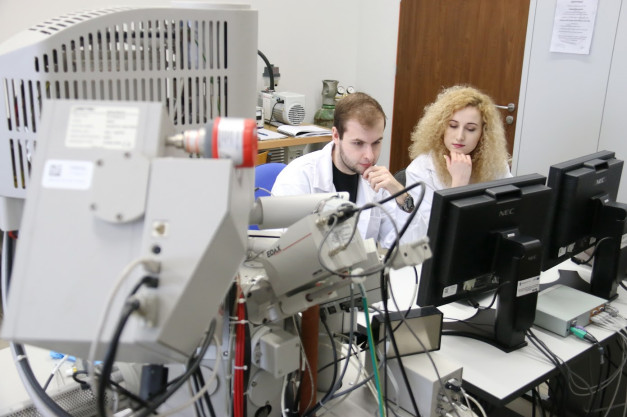 Dwóch naukowców w białych fartuchach pracuje przy skomplikowanym sprzęcie laboratoryjnym i monitorach.
