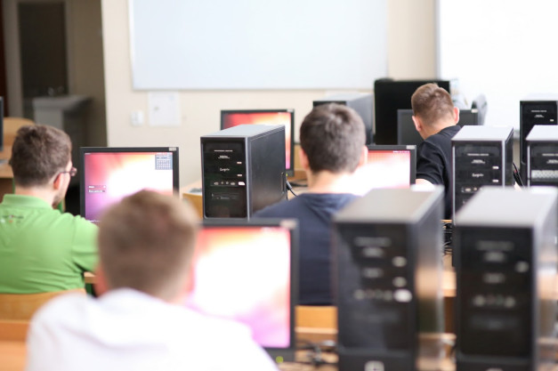 Osoby siedzą przy komputerach w sali lekcyjnej. Widać monitory, jednostki centralne i tablicę.