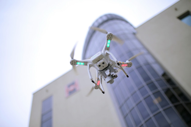 Biały dron z zielonymi i czerwonymi światłami leci przed nowoczesnym budynkiem.