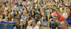 Widownia w sali wykładowej, głównie młodzi ludzie siedzą w rzędach. Kluczowe obiekty: ludzie, krzesła, sala wykładowa.
