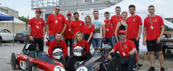 Grupa młodych ludzi w czerwonych koszulkach pozuje z dwoma bolidami. Kontekst: wydarzenie motoryzacyjne, studenci.