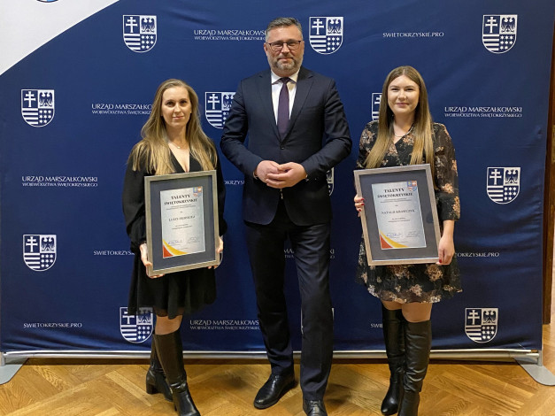 Trzy osoby stoją przed niebieskim banerem z herbami. Dwie kobiety trzymają dyplomy.