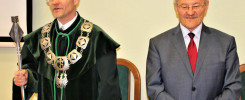 Dwóch mężczyzn w formalnych strojach stoi przy stole. Jeden w todze akademickiej, drugi w garniturze.