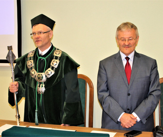 Dwóch mężczyzn w formalnych strojach stoi przy stole. Jeden w todze akademickiej, drugi w garniturze.