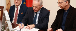 Trzech mężczyzn przy stole, jeden podpisuje dokument. W tle flagi.