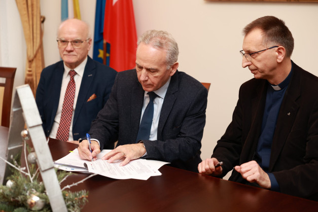 Trzech mężczyzn przy stole, jeden podpisuje dokument. W tle flagi.